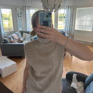 Beige stickad väst från Trendyol - Säljer en beige stickad väst från Trendyol med polokrage och snyggt kabelstickat mönster framtill. Västen är ärmlös och har ribbade kanter vid hals, ärmhål och nederkant. Perfekt att styla över en skjorta eller t-shirt för en chill look. En liten söm är lite lös på baksidan (se bild)