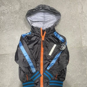 Svart vindjacka från Racoon  - Svart vindjacka från Racoon med huva, blå ränder på ärmarna och orange dragkedja framtill. Jackan har ribbade muddar i blått och svart, samt en vit dragkedja på bröstet. Märke med logga på vänster ärm. Perfekt för blåsiga dagar. Barnstorlek 98 
