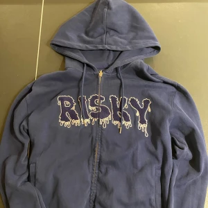 Mörkblå hoodie från Risky Universe - Säljer en mörkblå hoodie från Risky Universe med stor, broderad text 'RISKY' i vitt och blått på bröstet. Tröjan har huva med snörning, dragkedja och två fickor framtill. Grym streetwear-vibe och coolt drip-typsnitt på loggan. Storlek S, men passar mig som annars har storlek M.
