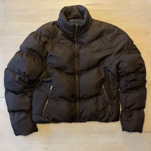 Brun pufferjacka från ASOS DESIGN - Brun pufferjacka från ASOS DESIGN med hög krage och två fickor med dragkedja. Jackan har en quiltad look och är tillverkad i syntetmaterial. Croppad passform och snörning nedtill. Perfekt för kalla dagar. Ena fickan är lite söndrig på insidan men märks knappt. Står 2XS på lappen men passar mer som XS-S.