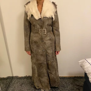 Lång pälsjacka från Najjar med bälte - Unik lång pälsjacka från Najjar, tillverkad i Italien. Jackan är brun med ett mönstrat skinnmaterial och har en stor vit pälsdetalj vid kragen. Den har bälte i midjan, knappar framtill och långa ärmar. Perfekt för dig som vill sticka ut med en retrovibe.