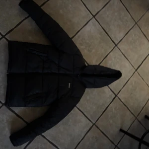 Svart pufferjacka från Hugo BOSS med huva - Svart pufferjacka från Hugo BOSS med huva och dragkedja framtill. Jackan har quiltad design, två sidofickor med dragkedja och en stilren look. Perfekt för kalla dagar och enkel att matcha med din vardagsstil.