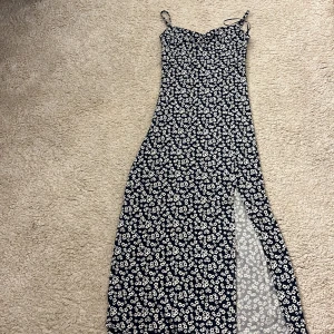 Marinblå blommig långklänning med slits - Snygg marinblå långklänning med vita blommor och smala axelband. Klänningen har en v-ringad överdel med rynkdetalj och en hög slits på sidan som ger en cool touch. Materialet känns lätt och flowy, perfekt för varma dagar.