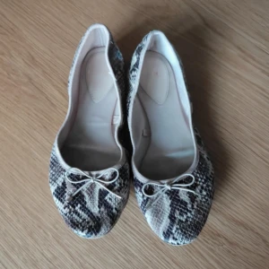 Ballerinaskor med ormmönster och rosett - Snygga ballerinaskor med ormmönstrad yta i beige och svart toner. Skorna har rund tå och en liten rosett framtill som detalj. Perfekta för dig som vill ha en cool och trendig look med platt sula och bekväm passform.