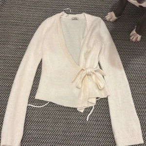 Vit omlottkofta från Gina Tricot - Superfin vit omlottkofta från Gina Tricot i stickat material. Koftan har långa ärmar, v-ringning och knyt i sidan som ger en snygg och feminin siluett. Perfekt att slänga på sig när det är lite kyligt ute. Ribbstickade detaljer vid muddar och nederkant.