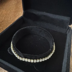 Guldigt tennisarmband med kristaller - Snyggt tennisarmband i guldton med glittrande vita kristaller längs hela bandet. Armbandet har en klassisk och stilren design med fyrkantiga infattningar och säker låsning. Perfekt för dig som vill ha något som verkligen blänker och drar blickarna till sig. Top kvalitet går att duscha med den undevik saltvatten för längre hållbarhet. 2,5mm tjock, 17cm lång. 