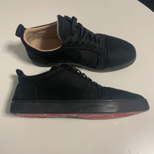 Svarta sneakers från Louboutin - Säljer ett par svarta sneakers från Louboutin med klassisk röd sula och snygg snörning. Skorna har rund tå, platt sula och är tillverkade i mocka med beige innersida. Perfekt för dig som gillar stilrena och exklusiva sneakers med en lyxig touch.