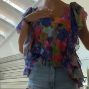 Blommig blus med volangärmar - Säljer en färgstark blommig blus med volangärmar. Toppen har en luftig och flowy passform med transparent material i lila, rosa, blå och gröna toner. Perfekt för dig som gillar att sticka ut med mönster och färg. Snygg att matcha med jeans eller kjol.