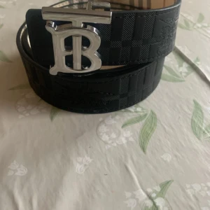 Burberry bälte med B-spänne - Säljer två Burberry-bälten med stort silverfärgat B-spänne. Ena bältet är svart med rutigt mönster och präglad Burberry-ryttare, det andra är beige med klassiskt rutigt mönster i rött, svart och vitt. Båda är breda och tillverkade i skinn med lyxig känsla.