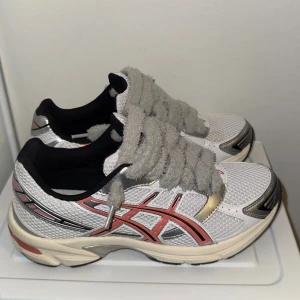 Vita Asics sneakers med fluffiga snören - Säljer ett par snygga Asics sneakers i vitt mesh med silvriga och röda detaljer. Skorna har fluffiga grå snören, svart foder och en platt sula. Perfekt för dig som gillar sportig stil och vill sticka ut med unika snören.