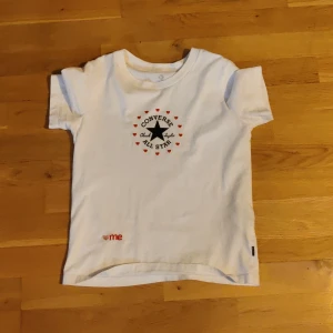 Vit Converse t-shirt med hjärtan  - Vit t-shirt från Converse med klassisk All Star-logga och svarta bokstäver. Runt loggan finns små röda hjärtan som ger en söt vibe. T-shirten har korta ärmar och är gjord i mjuk bomull. Perfekt för dig som gillar streetstyle och vill sticka ut lite extra.