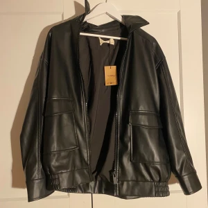 Svart skinnjacka från STR, storlek M - Säljer en svart skinnjacka från STR i storlek medium. Materialet är syntetiskt skinn och modellen är loose fit med elastiska detaljer vid nederkanten. 