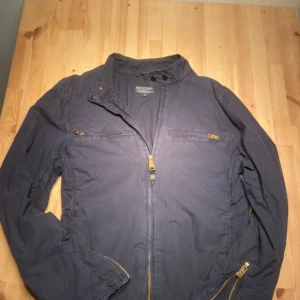 Mörkblå vindjacka Ralph Lauren - Snygg mörkblå vindjacka från Polo Ralph Lauren. Jackan har dragkedjor i guld, ståkrage med knappar och coolt tryck med amerikansk flagga och siffran 67 på ryggen. Tillverkad i 100% bomull, perfekt för dig som gillar street och sportig stil.