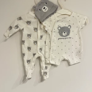 Baby rompers sets with hat for 0-1 months - New. Två par vita babybyxor med gulligt björnmönster i grått och texten 'I love Mum' och 'I love Dad'. Ena paret har grå stjärnor och en stor björn med texten 'growing every day'. Mjukt bomullsmaterial och tryckknappar framtill för enkel påklädning.
