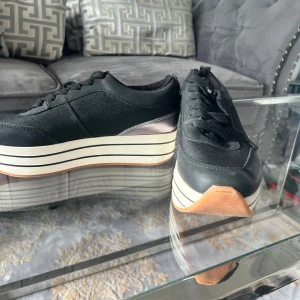 Svarta sneakers med platåsula från Zara - Säljer ett par svarta sneakers från Zara med cool platåsula i vitt med svarta ränder. Skorna har snörning och ovandel i textil och syntet. Snygg detalj med silvrig häl och gummisula i brunt. Perfekta för dig som vill ha lite extra höjd och stil.