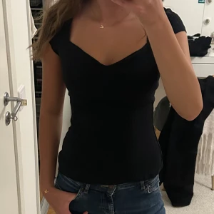 Svart ribbad topp med v-ringning - Snygg svart ribbad topp med v-ringad hals och korta ärmar. Figurnära passform som framhäver siluetten och passar perfekt till jeans eller kjol. Enkel men stilren design som är lätt att matcha.