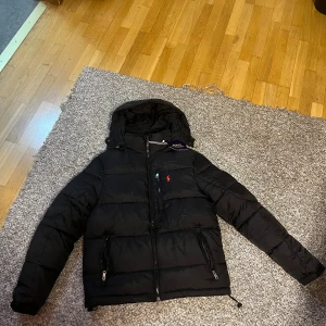 Svart pufferjacka från Polo Ralph Lauren - Svart pufferjacka från Polo Ralph Lauren med röd broderad logga på bröstet. Jackan har huva, två sidofickor med dragkedja och en innerficka. Materialet är syntet och jackan har quiltad design med normal passform. Perfekt för kalla dagar.