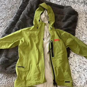 Vindjacka från Fjällräven - Snygg grön vindjacka från Fjällräven i storlek XXS. Sitter som en XS. Jackan har huva med snörning, vattentätt Hydratic-material och flera praktiska fickor med dragkedja. Perfekt för dig som gillar att vara ute och vill ha en cool och funktionell jacka.