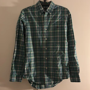 Rutig grön skjorta från Ralph Lauren - Snygg rutig skjorta från Ralph Lauren i grönt, blått och vitt med klassisk button-down krage och broderad logga på bröstet. Skjortan har långa ärmar och vita knappar framtill.