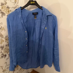 Blå skjorta från Polo Ralph Lauren - Blå skjorta från Polo Ralph Lauren i storlek L. Klassisk design med knappar framtill och lång ärm. Ikonisk gul broderad logga på bröstet. Skjortan är tillverkad i linne, vilket gör den lätt och skön att bära. Perfekt för dig som gillar stilrena och tidlösa plagg.