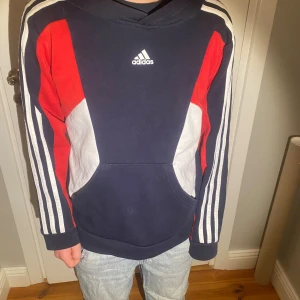 Adidas hoodie i blått, rött och vitt - Säljer en snygg Adidas hoodie med klassiska vita ränder längs ärmarna och Adidas-logga på bröstet. Tröjan är marinblå med röda och vita detaljer, har huva och känguruficka. Perfekt för dig som gillar sportig och avslappnad stil.