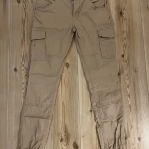 Beige cargopants från William Baxter - Säljer ett par beige cargopants från William Baxter i storlek S. Byxorna har flera praktiska fickor på sidorna och baktill, samt resår vid bensluten för en snygg och avslappnad look. Perfekta för dig som gillar streetstyle och funktionella detaljer.