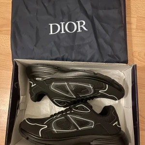  B30 svarta sneakers strl 43 - Säljer ett par svarta  B30 sneakers med coola silvriga detaljer och logga på både plös och häl. Skorna har en chunky platt sula, meshpaneler och snygga vita linjer som ger en futuristisk vibe. Perfekta för dig som vill sticka ut med lyxig streetstyle.