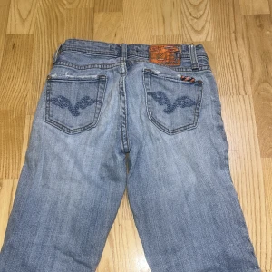 Vigoss ljusblå jeans med broderi - Säljer ett par ljusblå jeans från Vigoss med snygga broderade detaljer på bakfickorna och klassisk femficksdesign. Jeansen har normal passform och är tillverkade i mjukt denimtyg med orangea sömmar och logotyp på linningen. Står ej storlek men sitter bra på mig som har 32