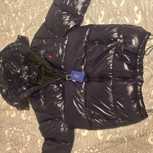 Mörkblå pufferjacka från Polo Ralph Lauren - Säljer en mörkblå pufferjacka från Polo Ralph Lauren med glansig finish och röd broderad logga på bröstet. Jackan har huva, dragkedjor på fickorna och är quiltad för extra värme. Perfekt för kalla dagar och har en riktigt snygg streetstil.