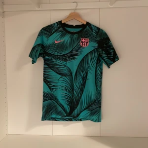 Barca tröja med palm tryck  - Barcelona Dri-Fit fotbollströja från Nike i XL, med turkos bas och svarta palmbladsmönster. Rosa Nike-logga och Barça-emblem på bröstet. Rund hals och korta ärmar, tillverkad i lätt polyester som andas. Perfekt för dig som vill sticka ut på planen.