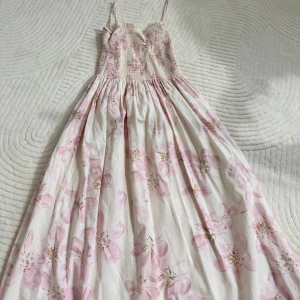 Lång blommig klänning med smock - Superfin långklänning med smala axelband och smockad överdel. Klänningen är vit med stora ljusrosa blommor och har en luftig, flowy kjol. Perfekt för varma dagar och ger en romantisk vibe med sitt blommiga mönster och mjuka färger.