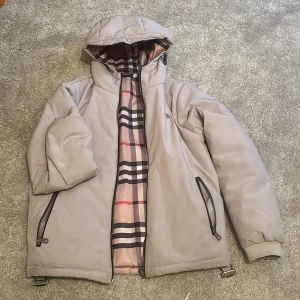 Beige pufferjacka från Burberry - Säljer en beige pufferjacka från Burberry med ikoniskt rutigt foder i rosa, rött, svart och vitt. Jackan har huva, två sidofickor med dragkedja och snygga Burberry-detaljer på dragkedjor och snörning. Perfekt för dig som vill ha en stilren och lyxig look.