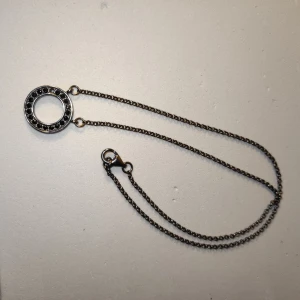 Mörk silverfärgad halsband med en rund cirkelberlock   - Snyggt halsband i silverfärgad metall med en rund cirkelberlock dekorerad med små svarta stenar runt kanten. Kedjan är tunn och har ett klassiskt lås. en stan har ramlat. 