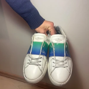 Valentino Garavani Open  - Snygga Valentino Garavani Open sneakers i vitt skinn med en bred, glansig panel i blått och grönt över mitten. Klassisk rund tå, platt sula och grå snörning. Ikoniska nitar baktill och logga på hälen. 