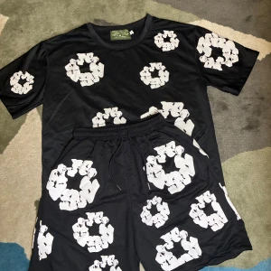 Denim Tears Set - Trendigt tvådelat set från Denim Tears, bestående av en svart t-shirt och matchande shorts med vit återvinningsinspirerad kransgrafik. Tillverkad i mjukt, lätt tyg för maximal komfort. Ett uttrycksfullt plagg med stark kulturell koppling och unik design från kollektionen African Diaspora Goods. Storlek: S.