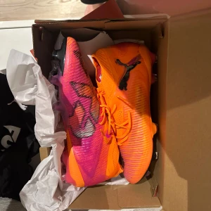 Puma orange/rosa fotbollsskor - Säljer ett par riktigt snygga Puma fotbollsskor i orange och rosa med svarta detaljer och logga. Skorna har snörning, platt sula med dobbar och en tight passform. Ovandelen är i syntetmaterial med coolt mönster och modern design, perfekta för dig som vill sticka ut på planen.