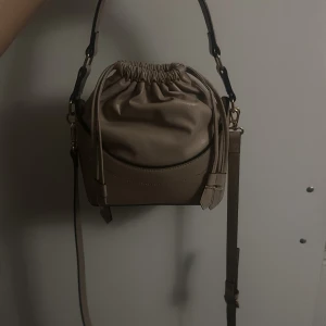 Beige axelväska med snörning - Snygg beige axelväska med cool snörning upptill och justerbar axelrem. Väskan har en mjuk, rundad form och är gjord i skinnimitation med guldfärgade detaljer. Perfekt storlek för det viktigaste och ger en chill vibe till din outfit. Väskan är från H&M🤎