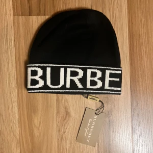 Svart Burberry mössa med logotyp - Svart mössa från Burberry med bred vit kant där BURBERRY är stickat i stora bokstäver runt om. Klassisk form och stilren design, perfekt för dig som gillar exklusiva märken. Materialet är stickat och känns mjukt och bekvämt mot huvudet.