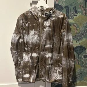 Woolrich vindjacka XL med mönster - Snygg vindjacka från Woolrich i storlek XL men sitter som L. Jackan är brun/grön med vitt abstrakt mönster, har huva och hel dragkedja framtill. Materialet är lätt och syntetiskt, perfekt för blåsiga dagar. Jackan är i perfekt skick, knappt använd. 