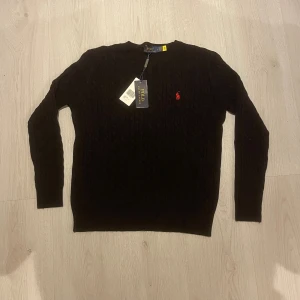Svart kabelstickad tröja Polo Ralph Lauren storlek M - Snabb leverans.  Storleken står det L men det kännes mer som M skulle jag säga Svart kabelstickad tröja från Polo Ralph Lauren med klassisk röd logobroderi på bröstet det är bra för en present eller för sig själv. Rund halsringning och ribbade muddar. Tröjan är långärmad och har en snygg, tidlös look som passar perfekt till jeans eller chinos.