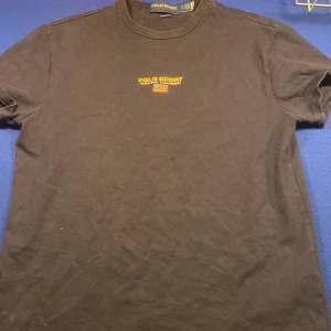 Svart Polo Sport t-shirt från Ralph Lauren - Svart t-shirt från Polo Sport Ralph Lauren med klassisk fit. T-shirten har korta ärmar och rund hals. På bröstet finns ett broderat Polo Sport-tryck i gult och en liten amerikansk flagga. Materialet är mjuk bomull, perfekt för en chill och stilren look.