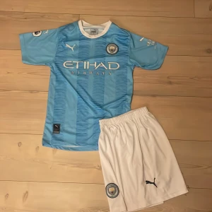 Manchester City Haaland matchtröja Puma - Manchester Citys matchtröja med Haaland 9 på ryggen. Ljusblå med vita detaljer, klubbmärke och Puma-logga. Tillverkad i lätt och ventilerande polyester. Säljes tillsammans med vita shorts med klubbmärke och Puma-logga.