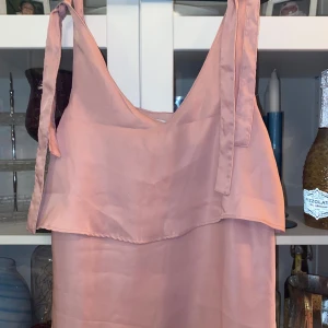 Blus - Säljer en ljusrosa blus med omlott-detalj och knytband på axlarna. Blus har ett lager över bysten som ger en cool och avslappnad look. Materialet är mjukt och har en lätt glans, perfekt för varma dagar.