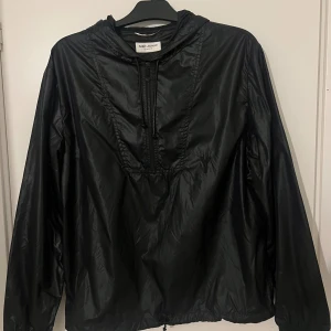 Yves Saint Laurent - vindjacka - Svart vindjacka från Saint Laurent med huva och halv dragkedja framtill. Jackan har en stilren design med Y-broderi på bröstet och justerbar snörning vid halsen. Inga skador eller fläck, såklart Äkta - vid önkan kan skicka detaljerade bild. Vid mer frågor är det bara att skriva.
