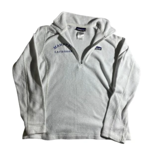 Vit fleece half zip från Patagonia - Vit fleece-tröja från Patagonia med broderad text 'MAHOPAC LACROSSE' på bröstet. Tröjan har half zip, hög krage och är långärmad. Tillverkad i mjuk polyesterfleece, perfekt för kyliga dagar.