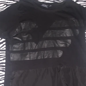 Svart t-shirt från Armani Jeans - Svart t-shirt från Armani Jeans med coola glansiga ränder framtill och diskret logga. Klassisk rund hals och korta ärmar. Materialet är mjuk bomull som känns skönt mot huden. Perfekt för dig som gillar stilrena plagg med en twist.