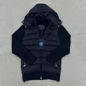 Marcel classic 1.5 Cardigan från Leon Marcel - Svart pufferjacka från Leon Marcel med stickade ärmar och huva. Jackan har dragkedja framtill och två sidofickor med dragkedja. Snygg, modern look med diskreta detaljer.