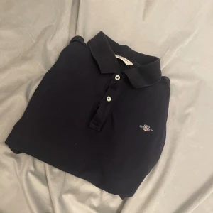 Svart pikétröja från GANT - Klassisk svart pikétröja från GANT med korta ärmar och krage. Tröjan har två vita knappar framtill och en broderad GANT-logga på bröstet. Materialet är mjukt och känns skönt mot huden, perfekt för en chill och stilren look.