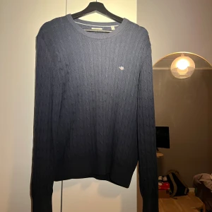 Mörkblå kabelstickad tröja från GANT - Snygg mörkblå kabelstickad tröja från GANT med rund hals och broderad logga på bröstet. Tröjan har långa ärmar och ribbade muddar vid hals, ärmslut och nederkant. Perfekt för dig som gillar klassisk och stilren look.