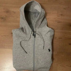Grå hoodie från Polo Ralph Lauren - Snygg grå hoodie från Polo Ralph Lauren med dragkedja och huva. Klassisk logga broderad på bröstet, två fickor framtill och justerbara snören i huvan. Tillverkad i mjuk bomullsmix, perfekt för chill dagar eller när du vill ha en clean look.